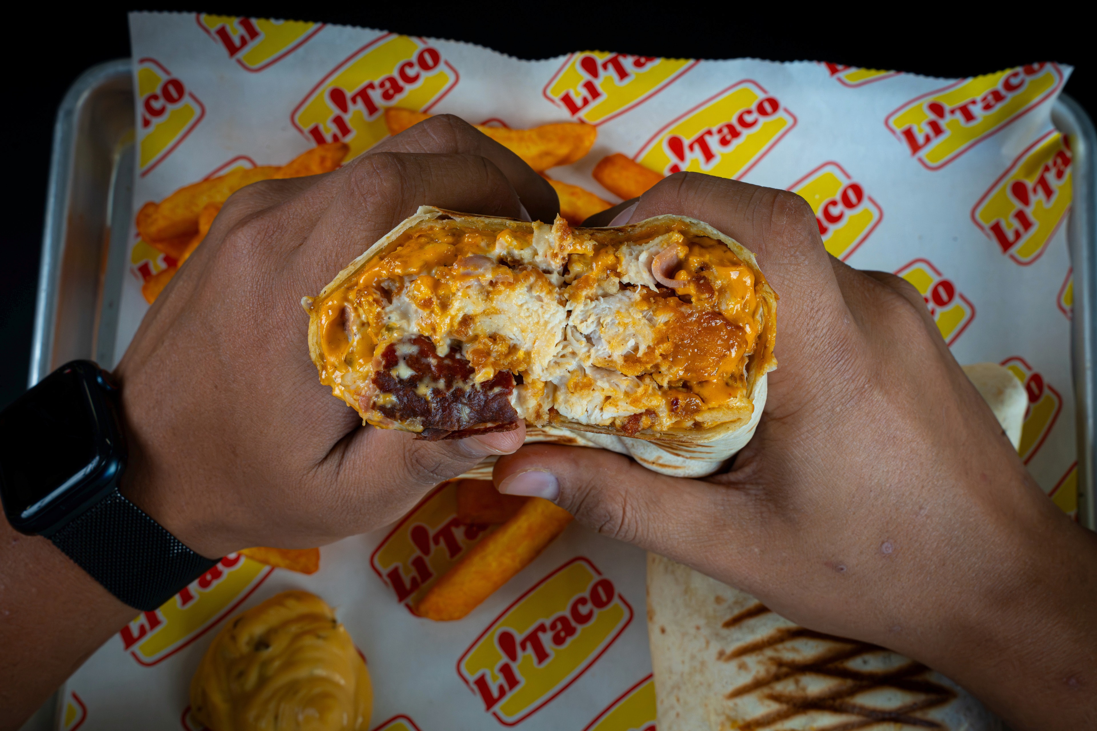 Dos manos abren un taco por la mitad sobre una bandeja con papel de LiTaco y patatas